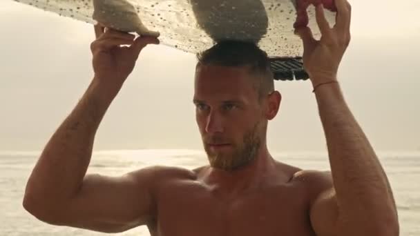 穏やかなひげを生やしたハンサムな男彼の頭の上にサーフボードを保持し 海の近くのビーチで離れて見て — ストック動画
