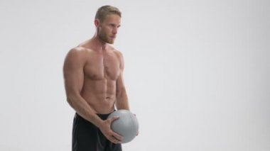 Odaklanmış genç, formda bir sporcu, beyaz arka planda izole edilmiş gri fitness topuyla egzersiz yapıyor.