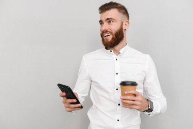 Çekici, yakışıklı bir iş adamının portresi gri arka planda duruyor, paket kahve içerken cep telefonu kullanıyor.