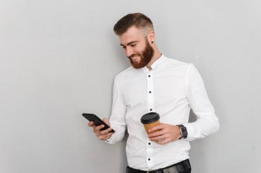 Çekici, yakışıklı bir iş adamının portresi gri arka planda duruyor, paket kahve içerken cep telefonu kullanıyor.