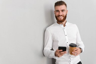 Çekici, yakışıklı bir iş adamının portresi gri arka planda duruyor, paket kahve içerken cep telefonu kullanıyor.