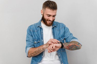 Gri arka planda dikilip, kol saatinin saatini kontrol eden, sıradan kıyafetler giyen yakışıklı bir gencin portresi.