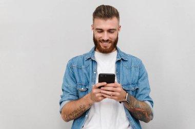 Gri arka planda izole bir şekilde duran, kablosuz kulaklıkla müzik dinleyen, elinde cep telefonu olan yakışıklı, güler yüzlü bir gencin portresi.