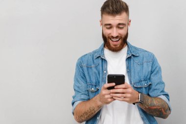 Gri arka planda izole bir şekilde duran, kablosuz kulaklıkla müzik dinleyen, elinde cep telefonu olan yakışıklı, güler yüzlü bir gencin portresi.