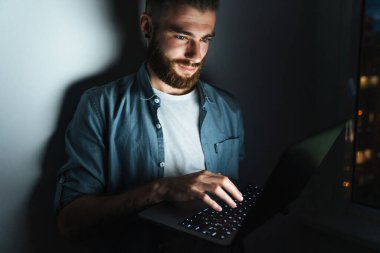 Diz üstü bilgisayarda çalışan kendine güveni tam genç bir adam geceleri evde duruyor, duvara yaslanıyor.