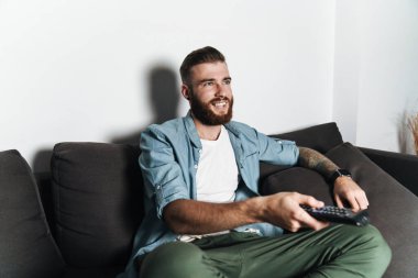 Çekici, gülümseyen, sakallı bir adam evdeki kanepede dinleniyor, televizyon izliyor, uzaktan kumanda tutuyor.