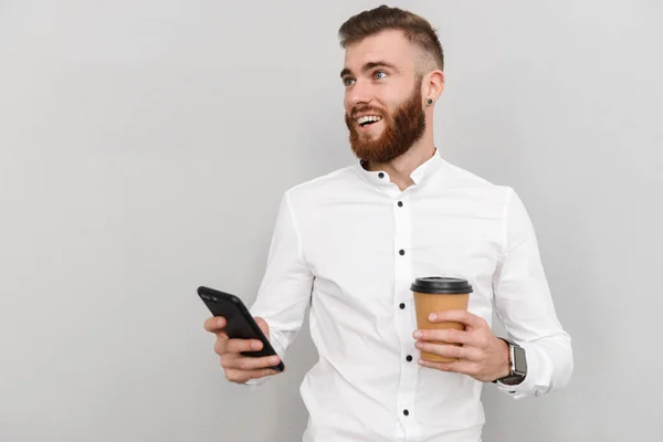 灰色の背景に立つ魅力的なハンサムな若いビジネスマンの肖像画 携帯電話を使用してテイクアウトコーヒーを飲みながら — ストック写真