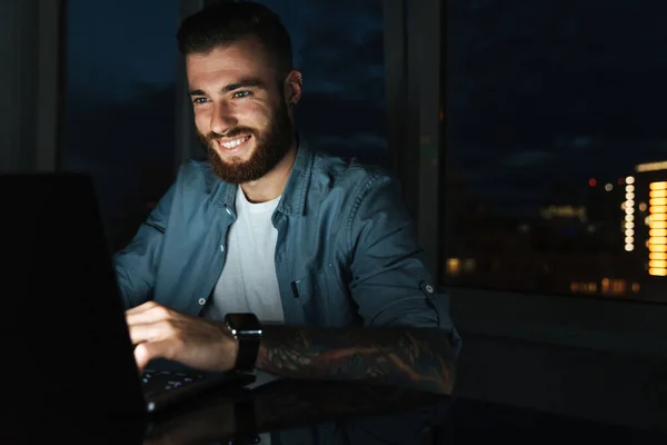 Usmívající Mladý Muž Pracující Notebooku Zatímco Sedí Noci Stolu — Stock fotografie