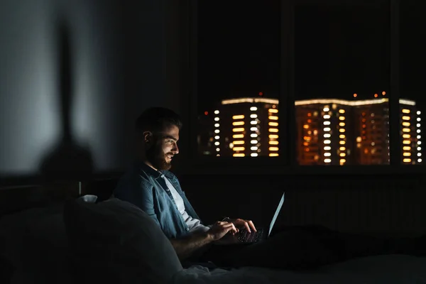 Smiling young bearded man laying on bed at home at night, working on laptop computer