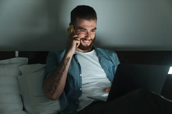 Усміхнений Молодий Бородатий Чоловік Лежить Ліжку Вдома Вночі Розмовляє Мобільному — стокове фото