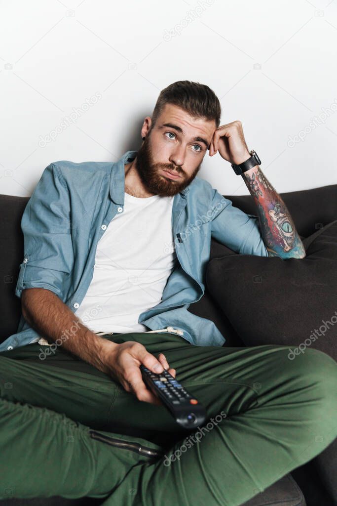 Attractive bored young bearded man relaxing on a couch at home, watching tv