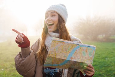 Şehir caddesinde elinde bir haritayla duran, kışlık ceket ve şapka giyen neşeli, genç bir kızın fotoğrafını çek.