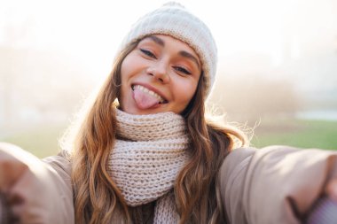 Şehir caddesinde durup selfie çeken, kışlık ceketi ve şapkası olan neşeli, genç bir kızın fotoğrafını çek.