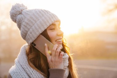 Şehir caddesinde dikilen, kışlık ceketi ve şapkası olan, cep telefonuyla konuşan neşeli, genç bir kızın yakın çekimi.