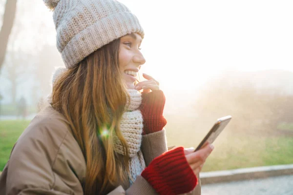 冬のジャケットを着た陽気な若い女の子と街の通りに立っている帽子のクローズアップ 携帯電話を使用して — ストック写真