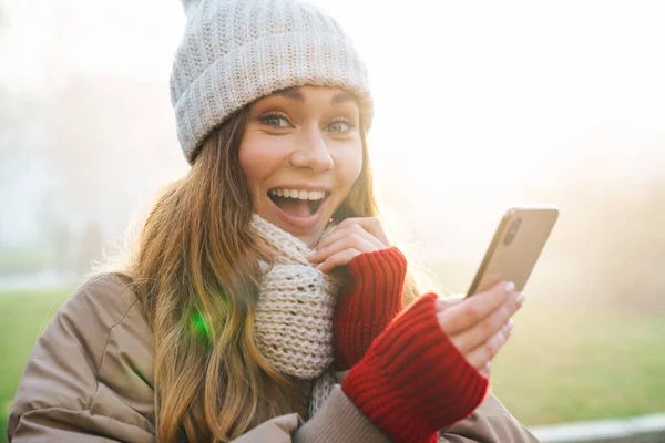 冬のジャケットを着た陽気な若い女の子と街の通りに立っている帽子のクローズアップ 携帯電話を使用して — ストック写真