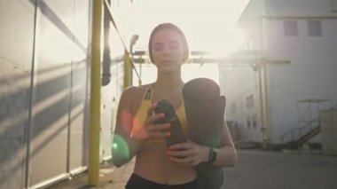 Spor giyimli sakin bir kadın yoga minderi tutarken dışarıda antrenman yapmak için sokağa çıkıyor.