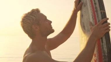 Sörf tahtasını temizleyen sakin kaslı sörfçünün gün batımında denizin kenarında dikilirken yan görüntüsü