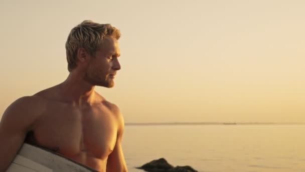 サーフボードを持って海の近くの夕日の上を歩くクールなひげそりの筋肉サーファー — ストック動画