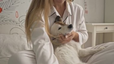 Beyaz pijama giyen çekici genç bir kadın yatak odasında yatarken bir köpeği tutuyor.