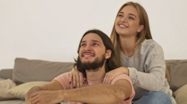 Mutlu, gülen bir çift, sıradan giyinmiş bir kadın ve erkek evdeki oturma odasında sarılırken televizyon izliyorlar.