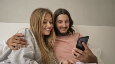 Mutlu genç bir çift cep telefonlarına bakarken evde yatakta uzanıyorlar.