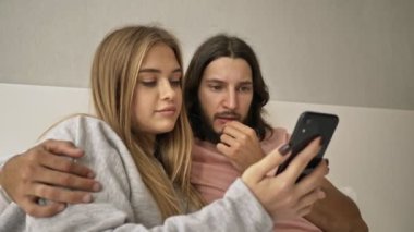 Kafası karışmış genç bir çift evde yatakta oturan bir cep telefonunu işaret ederken tartışıyor.