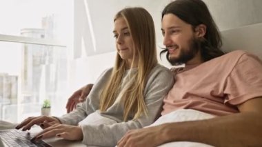 Mutlu, gülen genç bir çift evde yatarken dizüstü bilgisayarı birlikte kullanıyorlar.