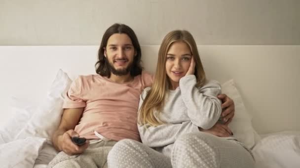 正の若いカップルの男性と女性は自宅で寝室のリモコンを使用してテレビを見ています — ストック動画