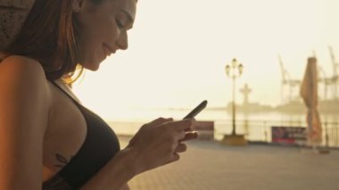 Neşeli, esmer ve sporcu bir kadının akıllı telefon kullanışının ve açık deniz kenarındaki bu anın tadını çıkarışının yan görüntüsü