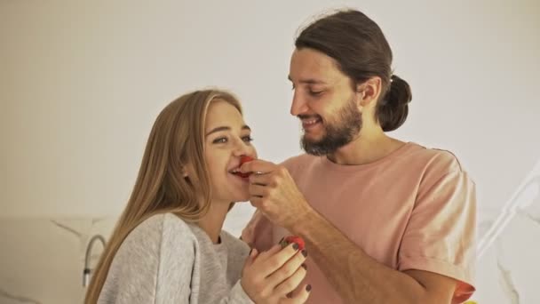 キッチンで料理をしながら 幸せな若いカップルがイチゴを食べ合っています — ストック動画