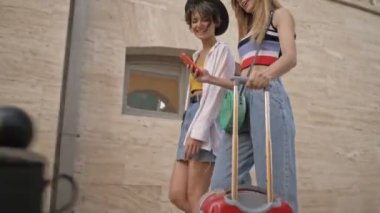 İki güzel turist kız akıllı telefon kullanıyor. Şehir caddesinde bavullarıyla dolaşıyorlar.