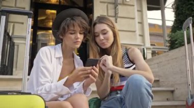 İki güzel turist kız akıllı telefon kullanıyor. Şehir caddesinde bavullarıyla geziyorlar.