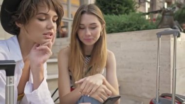İki gülümseyen duygusal turist kız şehir caddesinde akıllı telefon kullanırken konuşuyor.