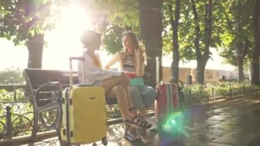 İki güzel genç turist kız bir şehir bulvarında bavullarıyla bankta otururken konuşuyor.
