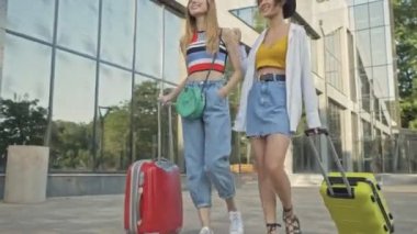 İki mutlu genç turist kız modern yüksek binaların yakınındaki bir şehirde bavullarıyla yürüyorlar.