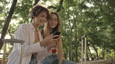 İki gülümseyen genç turist kız modern bir şehir parkında yürürken bir video görüşmesi yapıyorlar.