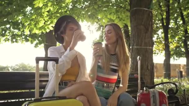 2人の笑顔の若い観光客の女の子がコーヒーを飲みながら街の大通りのスーツケースとベンチに座っています — ストック動画
