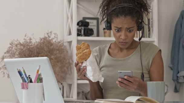 Σοκαρισμένη Όμορφη Αφρικανή Γυναίκα Ακουστικά Χρησιμοποιώντας Smartphone Και Τρώγοντας Σάντουιτς — Αρχείο Βίντεο