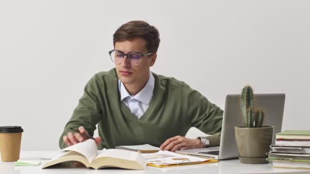 真剣な若い男学生はホワイトオフィスの机に座っている間に本を使って何かを書いている — ストック動画