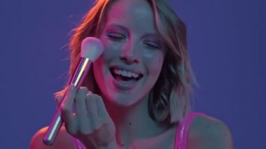 Mutlu genç bir kadının yakın plan görüntüsü mor neon arka planda izole edilmiş toz fırçası kullanarak makyaj yapıyor.