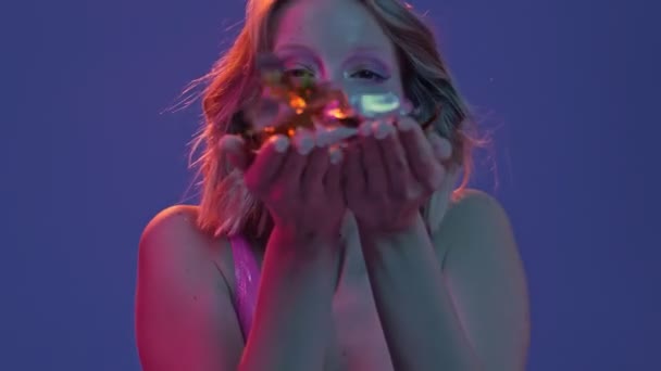 美しい笑顔の少女のクローズアップは 紫色のネオンの背景に隔離されたカメラにコンフェッティを吹いています — ストック動画