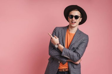 Çekici, ceketli ve şapkalı bir gencin portresi pembe arka planda izole bir şekilde duruyor ve fotokopi alanını işaret ediyor.