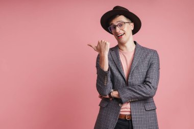 Çekici, ceketli ve şapkalı bir gencin portresi pembe arka planda izole bir şekilde duruyor ve fotokopi alanını işaret ediyor.