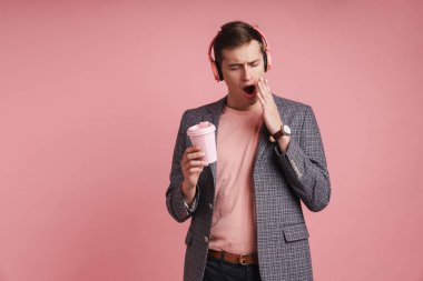 Pembe arka planda soyutlanmış, kablosuz kulaklıkla müzik dinleyen, elinde kahve fincanı tutan ceketli, yorgun ve çekici bir adamın portresi.