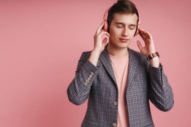 Pembe arka planda soyutlanmış, kablosuz kulaklıkla müzik dinleyen, gülümseyen, ceketli çekici bir adamın portresi.