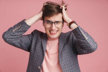 Pembe arka planda tek başına duran ve saçını koparan, ceketli, rahatsız bir genç adamın portresi.