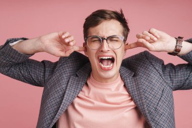 Pembe arka planda duran, kulaklarını tıkayan, ceketli, rahatsız genç ve çekici bir adamın portresi.