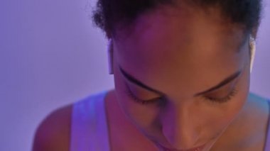 Kulaklıklı gülümseyen Afrikalı kadının yakın görüntüsü müzik dinliyor ve mor neon arka planda izole edilmiş anın tadını çıkarıyor.