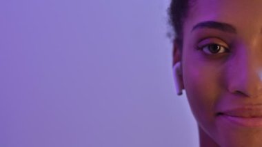 Kulaklıklı gülümseyen Afrikalı kadının yarısı müzik dinliyor ve mor neon arka planda izole edilmiş kameraya bakıyor.